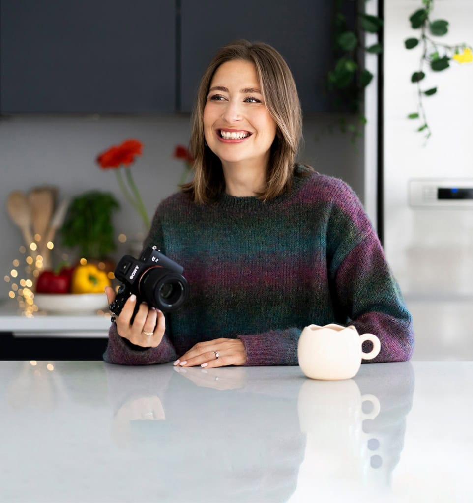 Photographe femme souriante tenant un appareil photo à la main dans une cuisine moderne, promouvant la nutrition et le bien-être avec une ambiance chaleureuse et naturelle.