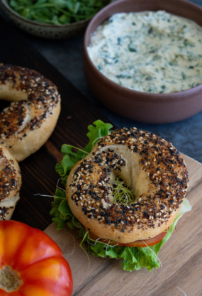 Bagel aux graines de sésame et de pavot avec légumes frais, servi avec une sauce crémeuse aux herbes, sur une planche en bois, mettant en avant une alimentation saine et savoureuse.