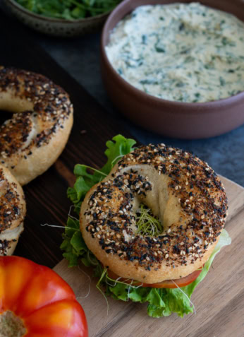 Bagel aux graines de sésame et de pavot avec légumes frais, servi avec une sauce crémeuse aux herbes, sur une planche en bois, mettant en avant une alimentation saine et savoureuse.