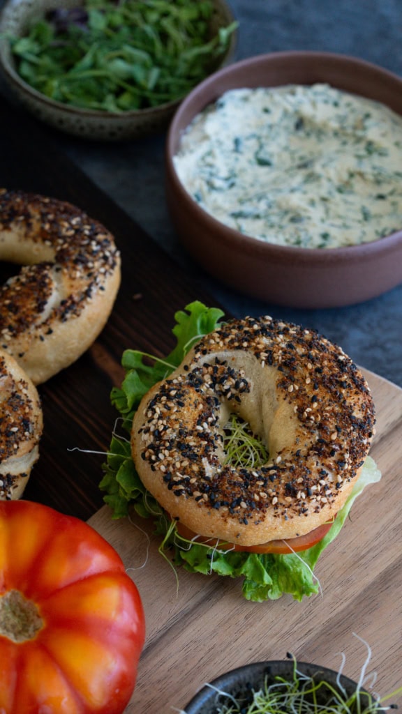 Bagel aux graines de sésame et de pavot avec légumes frais, servi avec une sauce crémeuse aux herbes, sur une planche en bois, mettant en avant une alimentation saine et savoureuse.