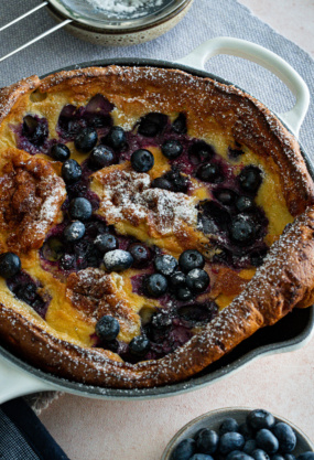 Gâteau aux myrtilles frais dans un plat de cuisson en céramique, avec un tamis à sucre et un bol de myrtilles fraîches, parfait pour une alimentation saine et une recette de dessert fruitée.