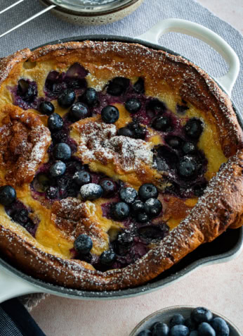 Gâteau aux myrtilles frais dans un plat de cuisson en céramique, avec un tamis à sucre et un bol de myrtilles fraîches, parfait pour une alimentation saine et une recette de dessert fruitée.