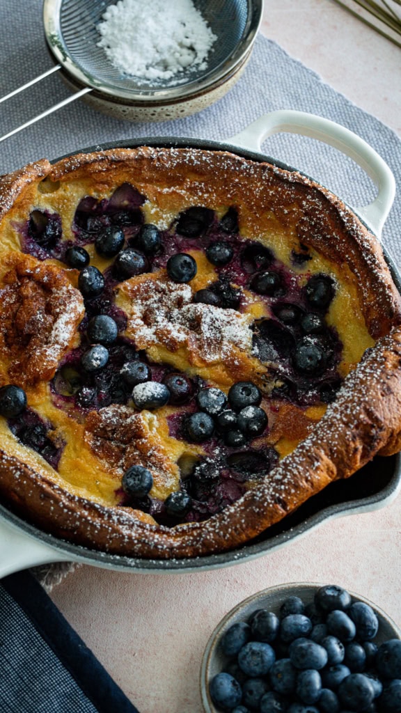 Gâteau aux myrtilles frais dans un plat de cuisson en céramique, avec un tamis à sucre et un bol de myrtilles fraîches, parfait pour une alimentation saine et une recette de dessert fruitée.