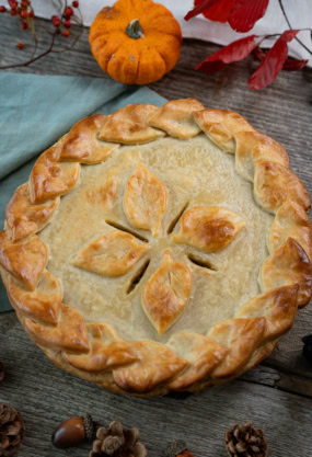 Galette des Rois à la Frangipane, pâtisserie traditionnelle de l'automne, décorée de feuilles en pâte feuilletée, entourée de décorations automnales comme des petits pumpkins, des pommes de pin et des châtaignes.