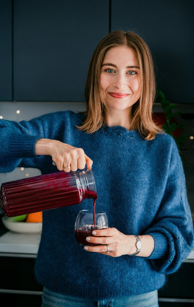 Amatrice souriante versant du jus de betterave dans un verre, dans une cuisine moderne, portant un pull bleu foncé, illustrant conseils en nutrition et alimentation saine.