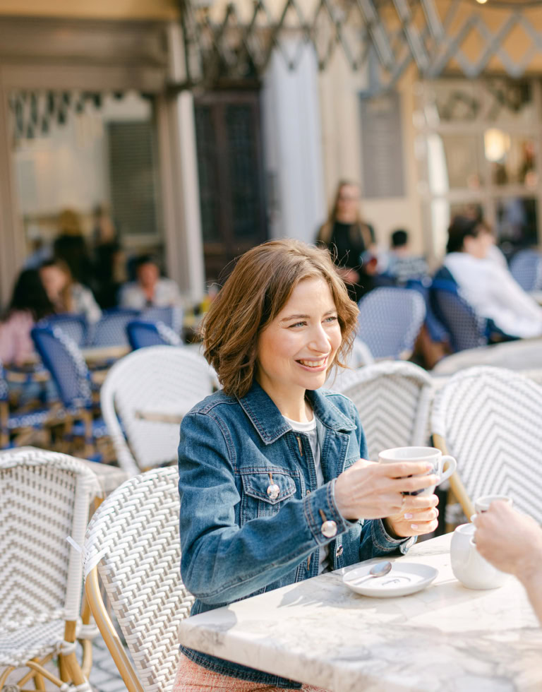 Jeune femme souriante dégustant un café dans un café en terrasse, ambiance chaleureuse et conviviale, avec d'autres clients en arrière-plan, ambiance urbaine et détendue.