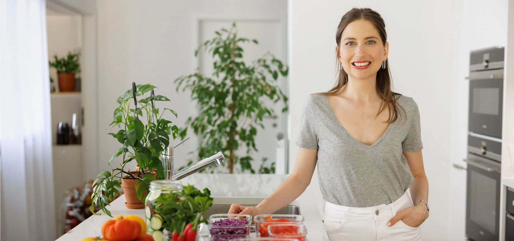 Femme souriante en cuisine avec des légumes frais et des plantes, promouvant une alimentation saine et équilibrée.